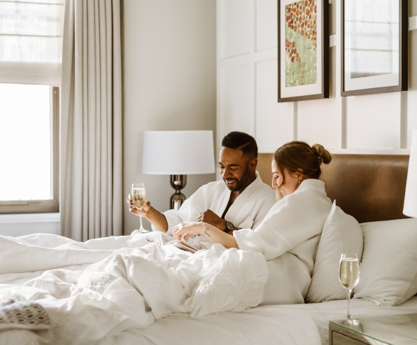 A couple in white bath robes drinking sparkling wine in a hotel bed.