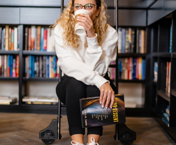 A woman sitting on a step ladder in a library drinking a coffee.